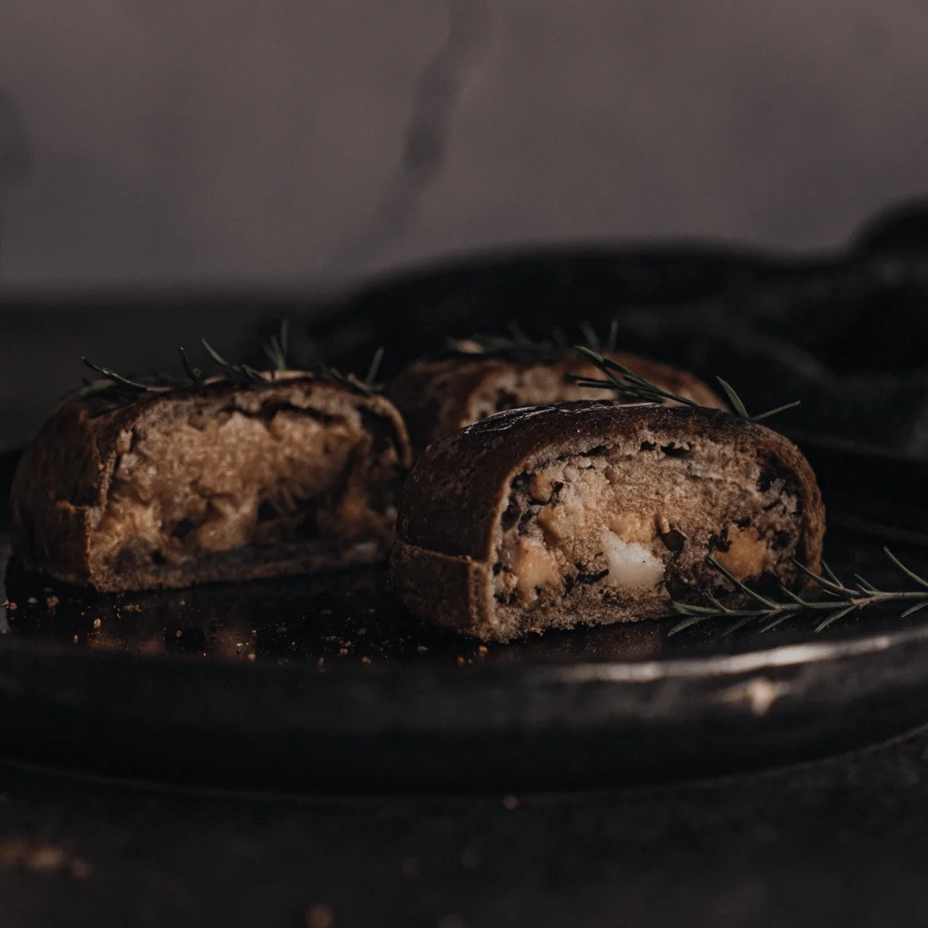 Dark, rustic pastries with a caramelized filling and a soft, textured crust, resembling caramel-infused rice cakes, served on a black plate with sprigs of rosemary.