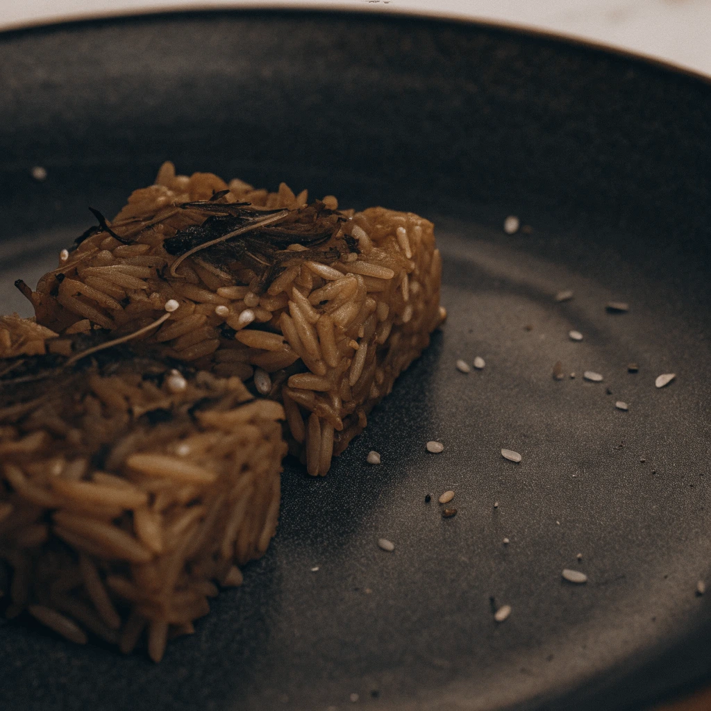 A close-up of two rectangular rice-based food portions on a dark plate, garnished with herbs and sprinkled with sesame seeds.