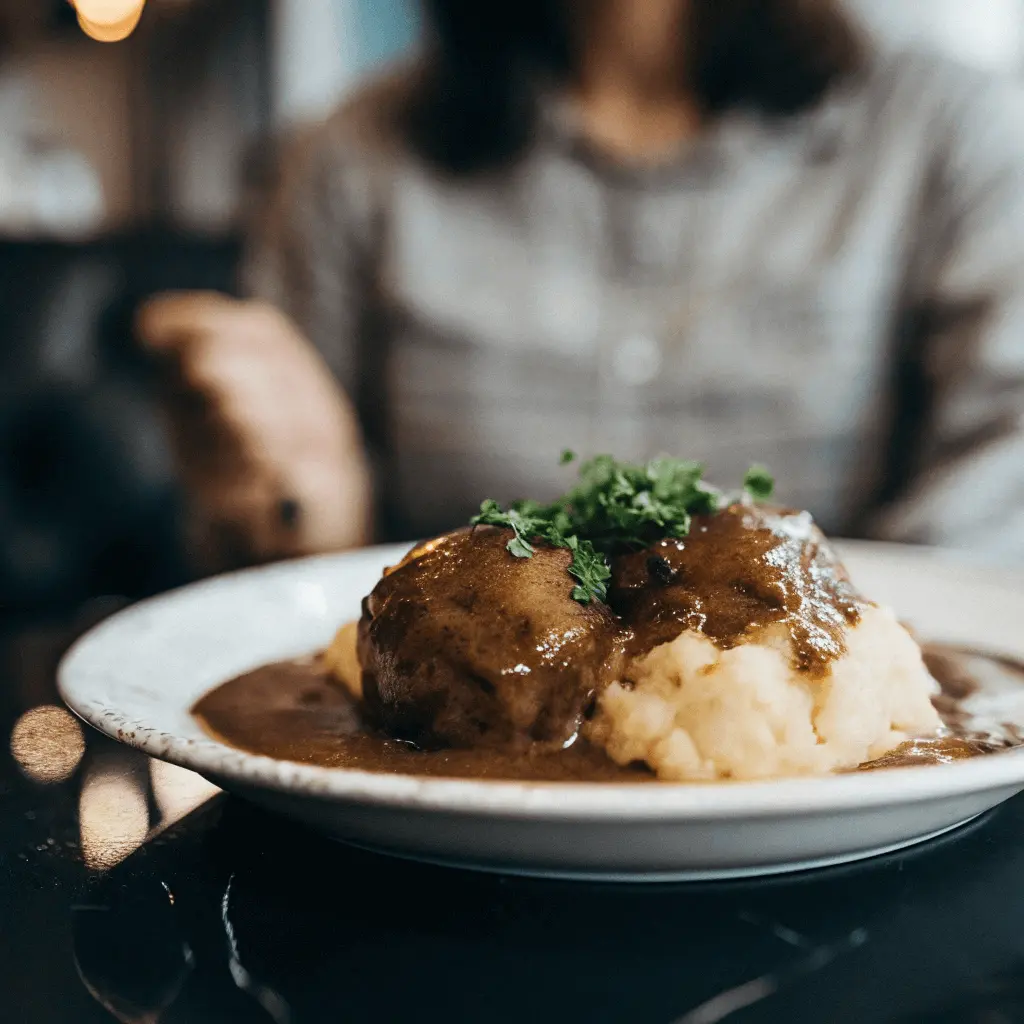 A plate of smothered potatoes with gravy, served alongside meatballs and garnished with fresh herbs.